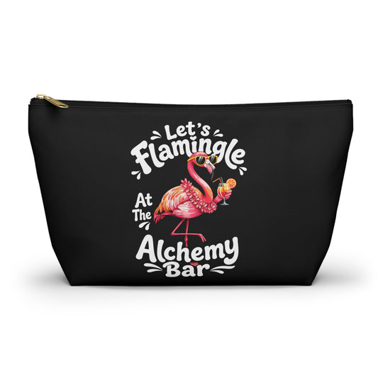 A black zippered cosmetic pouch featuring a colorful graphic of a pink flamingo holding a cocktail, surrounded by the text 'Let's Flamingle At The Alchemy Bar' in white and pink lettering.