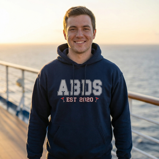 Man wearing a navy ABDS college-style hoodie with “EST 2020” and red martini accents while standing on a cruise ship deck at sunset.