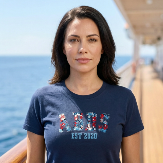 Woman wearing a navy ABDS floral collegiate-style tee with “EST 2020” while standing on a cruise ship deck with the ocean in the background.