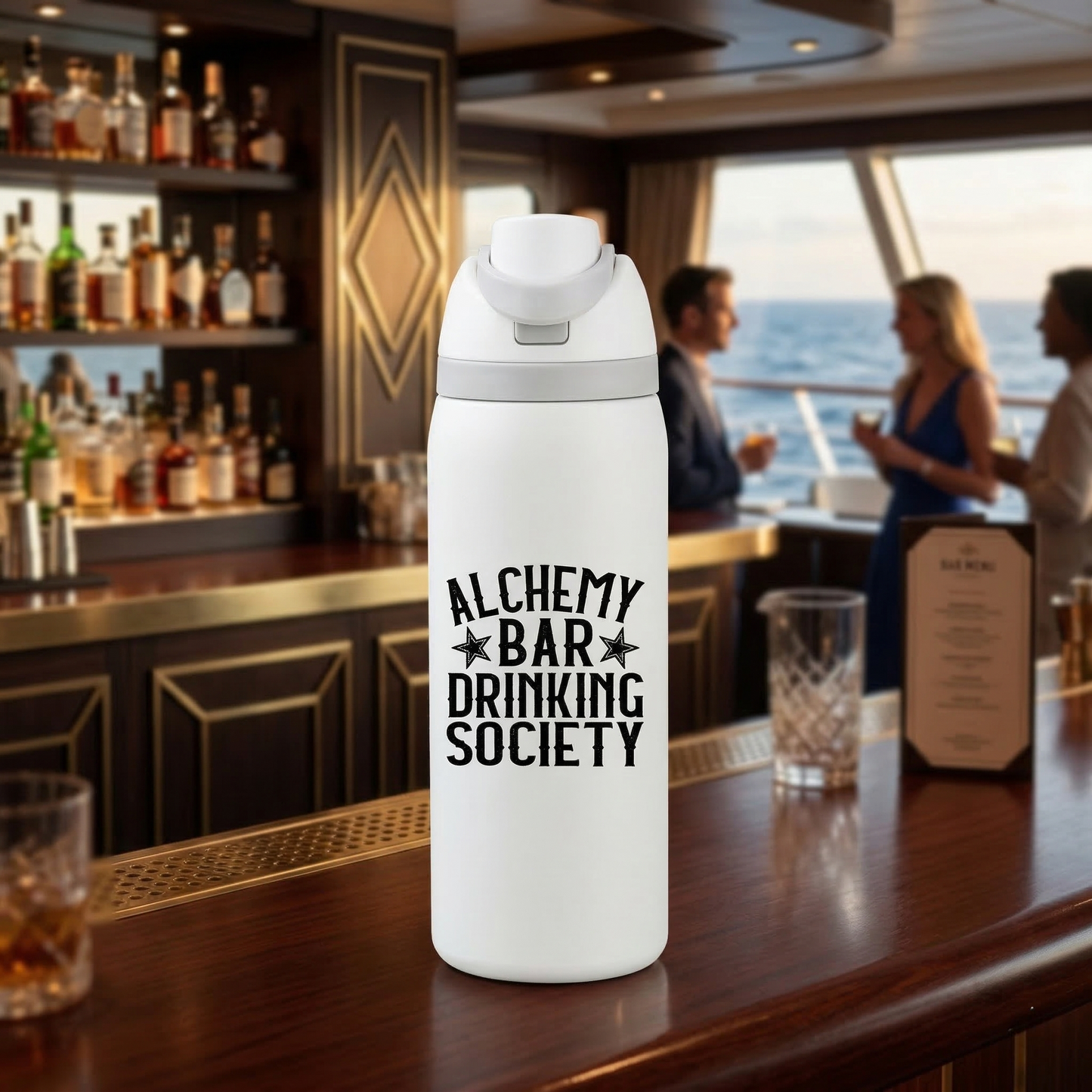 White insulated bottle with 'Alchemy Bar Drinking Society' text on a bar counter with people and drinks in the background.
